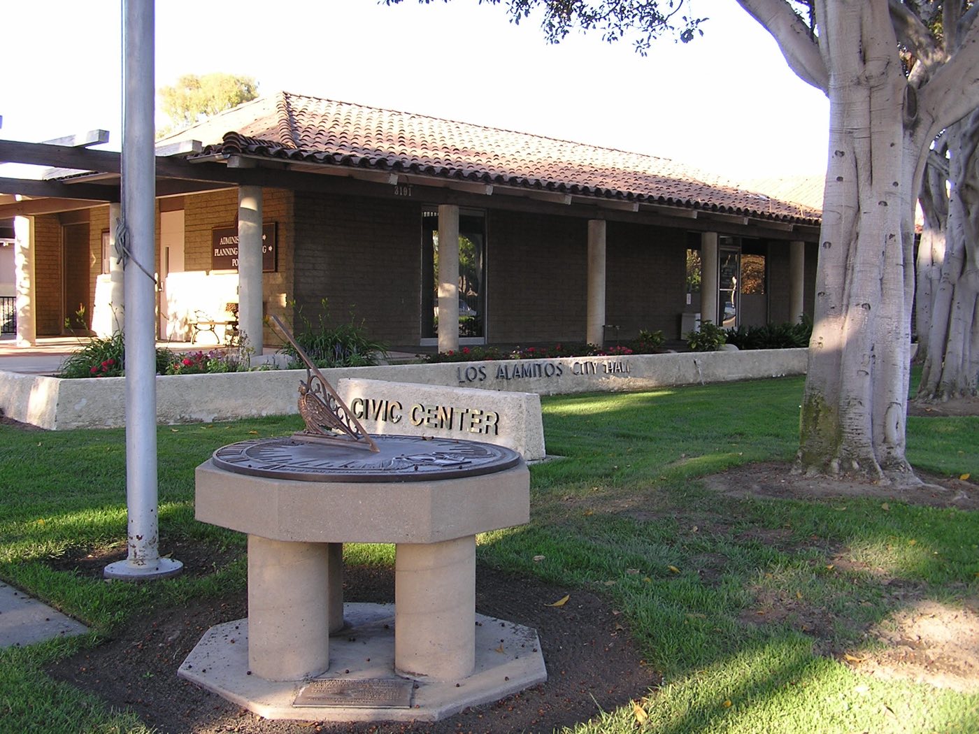 A bronze sundial on a lawn in front of a low, tile-roofed brick building. A concrete sign reads Civic Center, and lettering on the wall behind it reads Los Alamitos City Hall.