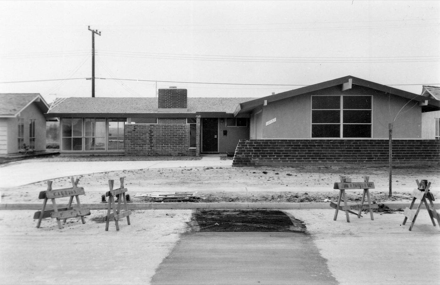 A black and white photograph from 1960. A newly built single-story tract house with a brick facade and a wide picture window stands on a bare dirt lot. Wooden contractor barricades line an unfinished street in the foreground.