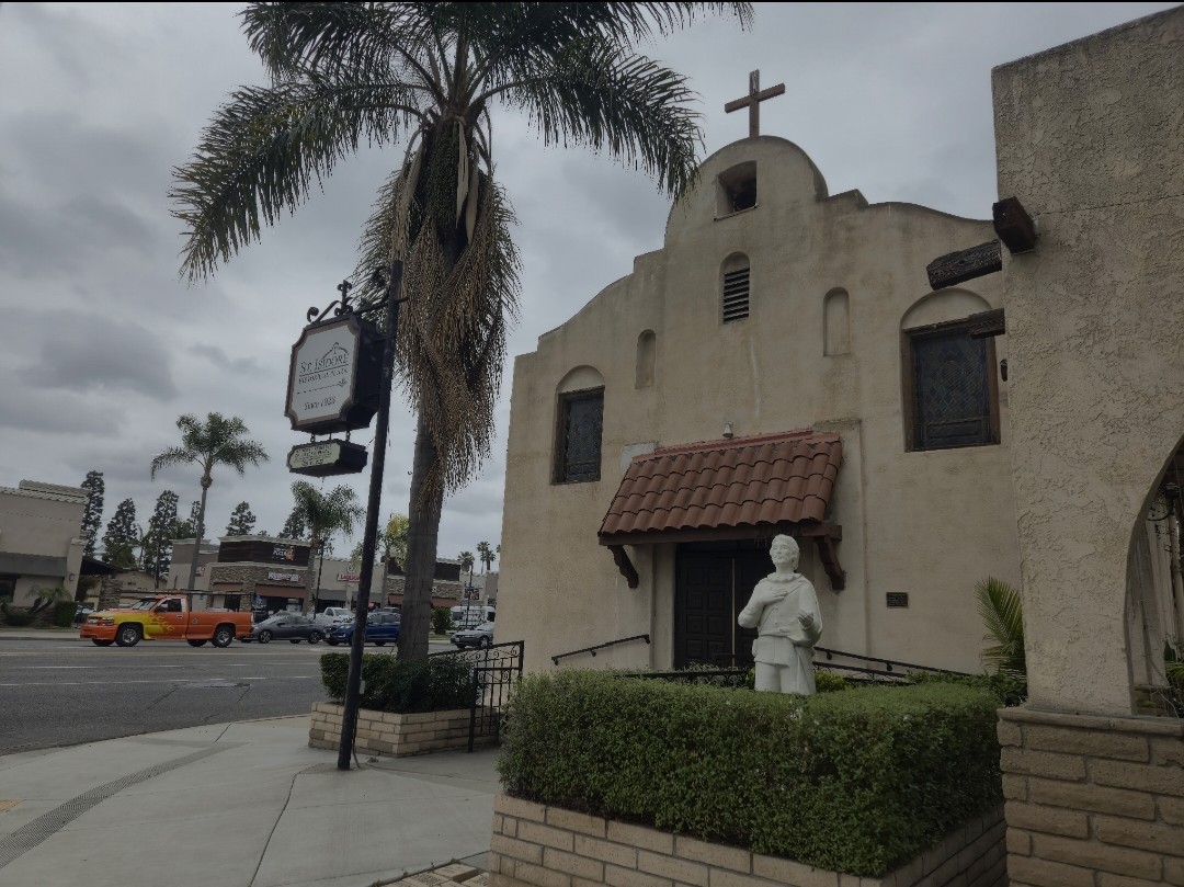 A white statue of a robed man stands in a low hedge in front of a cream-coloured mission-style church with a cross on its gable. A hanging sign and a palm tree stand at left, with Los Alamitos Boulevard and parked cars beyond.